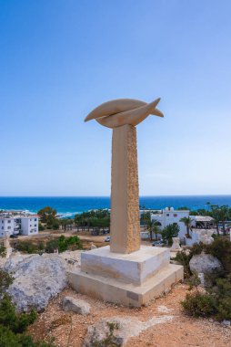 Akdeniz, beyaz binalar, yeşillikler ve palmiye ağaçları manzaralı kayalık bir tepenin üzerinde dokulu bir kaide üzerinde duran bir balık şekilli heykel..