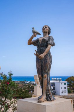 Kuşu tutan bronz bir kadın heykeli, açık mavi gökyüzüne, Akdeniz 'e, Kıbrıs' ta beyaz bir binaya ve yeşilliğe karşı duran bir kaide üzerinde duruyor..