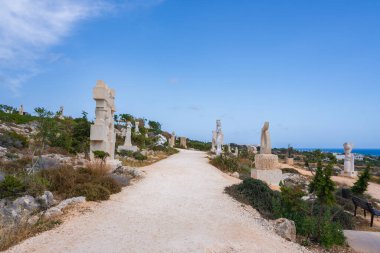Kıbrıs 'ta manzaralı bir yolda çeşitli taş heykeller, seyrek bitki örtüsü ve arka planda mavi gökyüzü ve deniz manzarası yer alıyor..