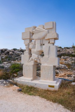Üzerinde insansı oymalar bulunan bir geometrik taş heykel kayalık, kuru çimenler ve Kıbrıs 'taki seyrek bitki örtüsüyle kurak bir zeminde..