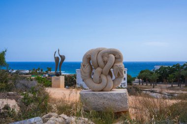 Kıbrıs Rum Kesimi 'nde bir tepede yılan ve soyut heykeller, yemyeşil palmiye ağaçları, beyaz binalar ve mavi deniz manzarası.