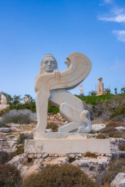 İnsan yüzüne, aslan vücuduna ve kanatlarına sahip bir sfenks, Kıbrıs 'taki kayalık bir yamaçta, seyrek bitki örtüsü ve diğer heykellerle çevrili..
