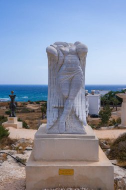 Heykeller, bitki örtüsü ve yakınlarındaki modern binalarla Akdeniz yakınlarında açık havada sergilenen plaketli bir kaidedeki soyut beyaz mermer heykel..