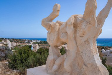 Yeşillik, uzak heykeller ve Kıbrıs 'taki derin mavi Akdeniz' in bulunduğu kıyı manzarasında soyut taş heykel.