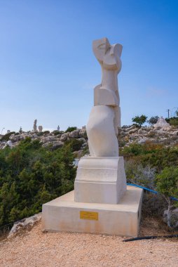 Kayalık arazi, seyrek yeşillik ve açık mavi gökyüzü altında ek heykeller ile çevrili, bir tabela üzerinde modern soyut taş heykel.