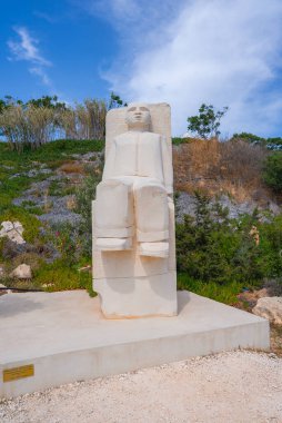 Beton bir platformun üzerinde oturan büyük beyaz taş bir heykel, parçalı bulutlu mavi gökyüzünün altında çalıları olan doğal bir yamaca karşı kurulmuş..