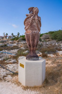Beton bir kaideye oyulmuş ahtapot heykeli, açık bir gökyüzünün altında kayalık, kurak, seyrek bitki örtüsü ve palmiye ağaçları olan bir arazide sergileniyor..