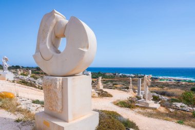 Kıbrıs kıyı şeridi yakınlarındaki bir heykel parkında kumlu bir yol boyunca çağdaş taş heykeller, canlı bir mavi deniz ve arka planda açık bir gökyüzü yer alıyor..