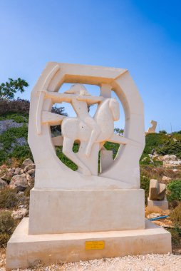 Dairesel bir çerçeve içinde kayalık arazi, seyrek yeşillik ve açık mavi gökyüzü arasında bir kaide üzerine kurulu bir at ve binicinin beyaz taş heykeli.