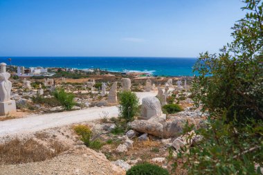 Kıbrıs 'ta kayalık arazide taş heykeller, mavi denize giden kirli bir yol ve ufukta görünen küçük binalar olan bir kıyı manzarası..