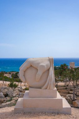 Bir küreyi tutan el şeklindeki heykel Akdeniz yakınlarında kayalık arazi ve seyrek bitki örtüsüyle çevrili bir taş kaide üzerinde duruyor..