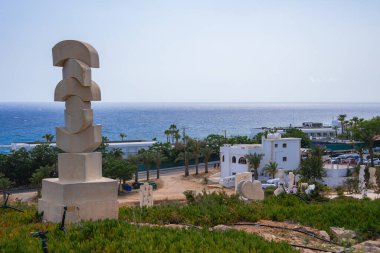 Kıbrıs 'ta bir tepede modern bir soyut taş heykel, beyaz bir Akdeniz tarzı bina, palmiye ağaçları ve arka planda okyanus.