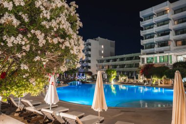Işıklı bir yüzme havuzu, şezlonglar, şemsiyeler ve ön planda beyaz çiçekleri olan bir ağacın yer aldığı bir Kıbrıs otelinin gece manzarası.