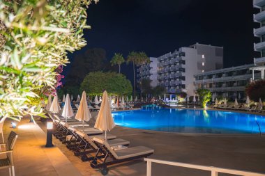 Kıbrıs 'ta ışıklandırılmış yüzme havuzu, şezlonglar, şemsiyeler, palmiye ağaçları ve arka planda modern otel binaları bulunan bir tatil köyü..
