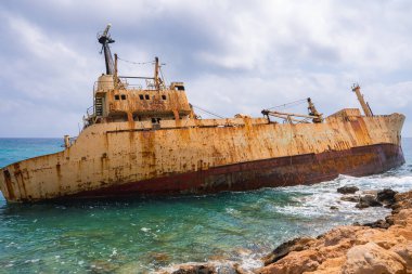 Edro III 'ün gemi enkazı, bulutlu bir gökyüzünün altında canlı turkuaz sulara dayanan paslı gövdesiyle Kıbrıs, Paphos' un kayalık kıyıları yakınında bulunuyor..