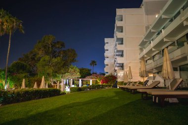 Balkonları olan modern beyaz bir otel açık bir gece gökyüzünün altında şezlongları, şemsiyeleri, palmiyeleri ve canlı çiçekleri olan çimenli bir alanın yanında duruyor..