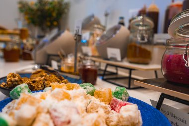 Bir tabak pudralı Türk lezzeti büfedeki baharat ve sos kavanozlarıyla çevrili. Arka planda bulanık modern yemek alanı.