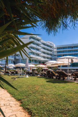 Kıbrıs 'ta balkonları, şezlongları, şemsiyeleri ve açık hava altında çimenli bir alanda güneşli hava altında palmiyeleri olan modern çok katlı bir otel..