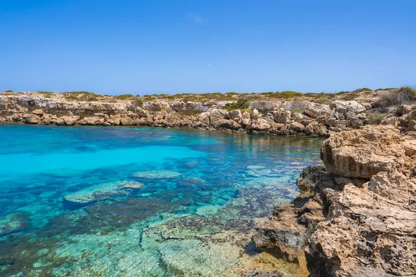 Kristal berrak turkuaz sular, engebeli bir kıyı şeridinde, açık renkli oluşumları ve mavi gökyüzünün altında alçak bitki örtüsü olan su altındaki kayaları ortaya çıkarıyor..
