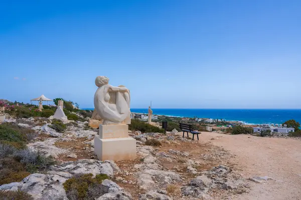 Kıbrıs 'ta kayalık bir tepede arpla oturan beyaz mermer bir heykel, Akdeniz' e tepeden bakan berrak bir gökyüzü ve yemyeşil.