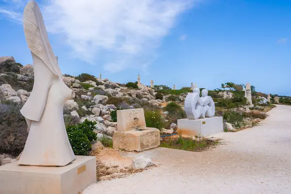 Kıbrıs 'taki bir heykel parkında, kayalık arazi ve açık mavi gökyüzü ile çevrili, toprak bir patika boyunca uzanan kaidedeki modern taş heykeller yer alıyor..