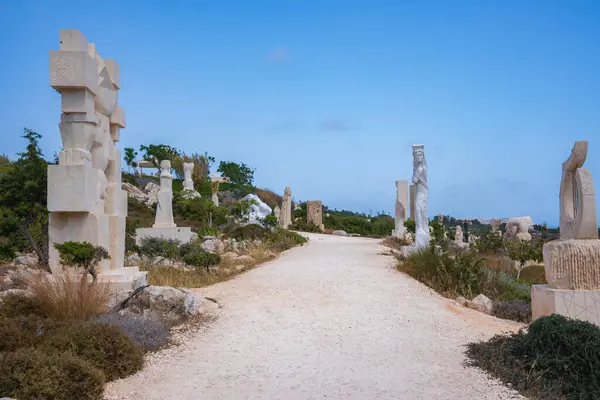 Kıbrıs 'taki sakin bir patikada, açık ve kurak bir arazide, açık bir gökyüzü altında seyrek bitki örtüsü ve uzak tepeler bulunan çeşitli taş heykeller yer alıyor..
