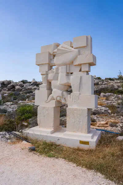 Üzerinde insansı oymalar bulunan bir geometrik taş heykel kayalık, kuru çimenler ve Kıbrıs 'taki seyrek bitki örtüsüyle kurak bir zeminde..