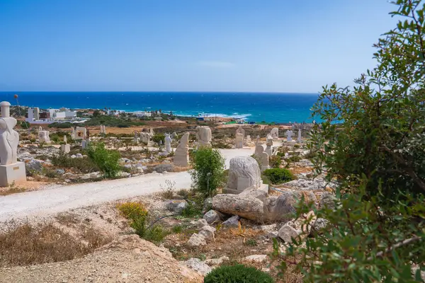 Kıbrıs 'ta kayalık arazide taş heykeller, mavi denize giden kirli bir yol ve ufukta görünen küçük binalar olan bir kıyı manzarası..