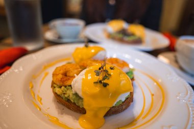 Avokado soslu Benedict yumurtası, haşlanmış yumurta, Hollandez sosu ve mikroyeşillik. Arka planda benzer bir yemek, kahve ve su var..