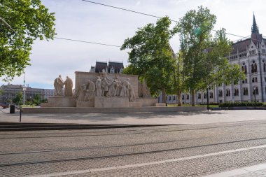 Arka planda ağaçlar, tramvay rayları ve Macar Parlamento Binası tarafından çerçevelenmiş, halka açık bir meydanda ayrıntılı heykeller bulunan önemli taş heykel.