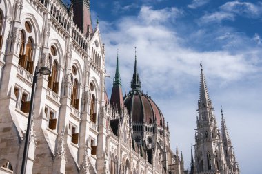 Budapeşte 'deki Macar Parlamento Binası' nda dağınık bulutlu parlak mavi gökyüzüne karşı dizilmiş kemerler, kuleler ve merkezi bir kubbe yer alıyor..