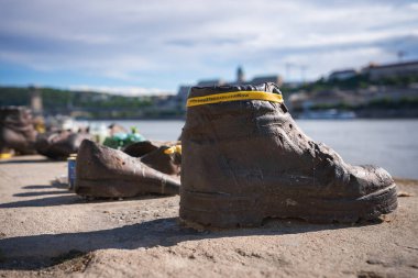 Budapeşte, Macaristan 'daki Yahudi Soykırımı Anıtı' nda sarı bantlı bronz ayakkabı heykeli. Tuna Nehri ve Buda Şatosu parçalı bulutlu gökyüzünün altında görünür.