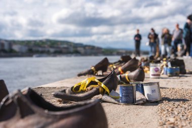 Demir ayakkabılar Macaristan 'ın Budapeşte kentindeki Tuna Nehri' nde sarı kurdele ve teneke kutularla hizalanır. Bulanık şekiller ve şehir manzarası parçalı bulutlu bir gökyüzünün altında belirir.