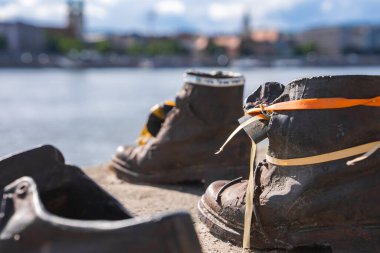 Paslı demir ayakkabılar ve turuncu ve sarı kurdelelerle Tuna Nehri anıtındaki Ayakkabılar, arka planda Tuna Nehri ve şehir yapıları.