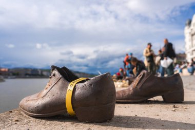 Bronz ayakkabılar Macaristan 'ın Budapeşte kentindeki Tuna Nehri kıyısında soykırım kurbanlarını onurlandırıyor. Sarı bir şerit görülebilir, içinde insanlar ve manzarası var..