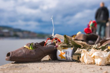 Bronze shoes on the Danube Bank in Budapest, Hungary, adorned with flowers and a candle. The Danube River and cityscape appear in the background.