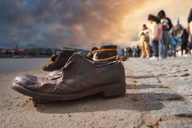 Soykırım anıtının bronz ayakkabıları Budapeşte 'deki Tuna Nehri kıyısı, bir grup insan ve arka planda sıcak bir günbatımı gökyüzü..