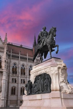 Ferenc Rakoczi II 'nin süslemeli bir heykeli arkasında pembe ve mor bir gökyüzü bulunan neo Gotik Macar Parlamento Binası önünde duruyor..