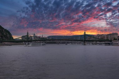 Yeşil çelik Özgürlük Köprüsü Tuna Nehri boyunca canlı bir günbatımı altında uzanır. Gellert Tepesi ve tarihi binalar şehrin zeminini oluşturuyor..