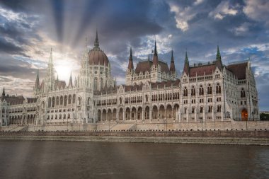 Budapeşte 'deki Tuna Nehri' nin yanında yer alan neo Gotik Macar Parlamentosu Binası 'nın önünde bulutların arasından süzülen güneş ışığı ve ön plandaki sakin sular yer alıyor..