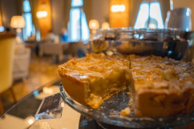 Kısmen dilimlenmiş altın hamurlu ve cam bir tabakta parıltılı bir turta, sıcak ışıklandırması ve kemerli pencereleri olan butik bir otelin iç kısımlarına kurulmuş..