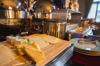 Ahşap bir tahtanın üzerinde zarif bir peynir büfesi. Taze otlar, metalik servis kubbeleri ve iyi aydınlatılmış bir butik otel yemek alanında çatal bıçak takımı..