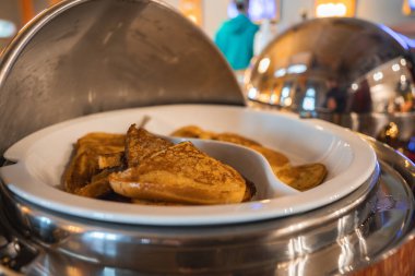 Altından kahverengi krepleri paslanmaz çelik bir aşındırma kabında, sıcak ışıklandırmalı, yansıtıcı yüzeyli ve arka planda yeşil kapüşonlu kapüşonlu bir kişi.