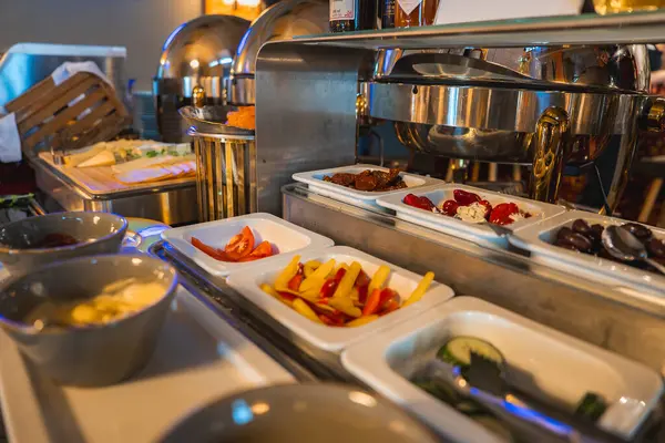 Üst düzey bir büfede taze sebzeler, doldurulmuş biberler ve beyaz tepsilerde zeytinler, metalik aşındırıcı kaplar ve arka planda sıcak ışıklandırma yer alıyor..