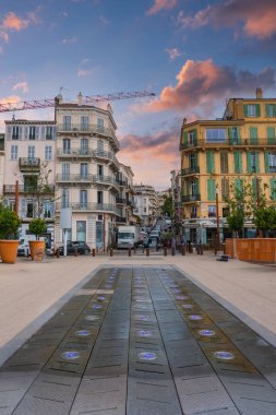 Fransa 'nın Cannes kentindeki bir sokakta dairesel ışıklar, pastel Fransız tarzı binalar, saksı ağaçları ve arka planda bir vinç yer alıyor..