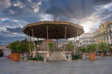 Fransa, Cannes 'da bir kaldırım taşı plazasında karmaşık demir işçiliğiyle süslü bir çardak. Yeşillik, klasik binalar ve kırmızı kiremitli çatısı olan bir kilise..