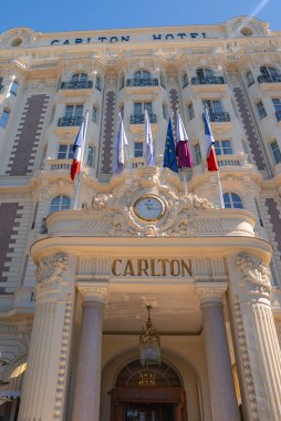 Fransa 'nın Cannes kentindeki Carlton Otel' de dekoratif sütunlar, merkezi bir saat ve sergilenen birden fazla bayrakla süslü bir krem rengi cephe bulunuyor..