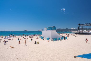 Cannes, Fransa 'da güneşli bir sahil, Cinema de la Plage, dinlenen insanlar, turkuaz sular, demirlemiş yatlar, ve uzak manzaralı tepeler.
