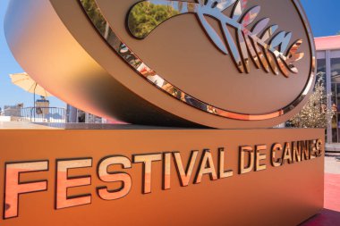 Altın Cannes Festivali logosu, ağaçlar ve açık hava oturma yerleri ile işaretlenir. Arka plandaki kırmızı halı Fransa 'nın Cannes şehrinde güneşli bir gün olduğunu gösteriyor..