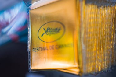 Cannes Festivali metni ve simgesel palmiye yaprağı logosunun yansıtıcı yüzeyi vurgulayan bulanık bir arkaplan içeren altın pakete yakın plan.